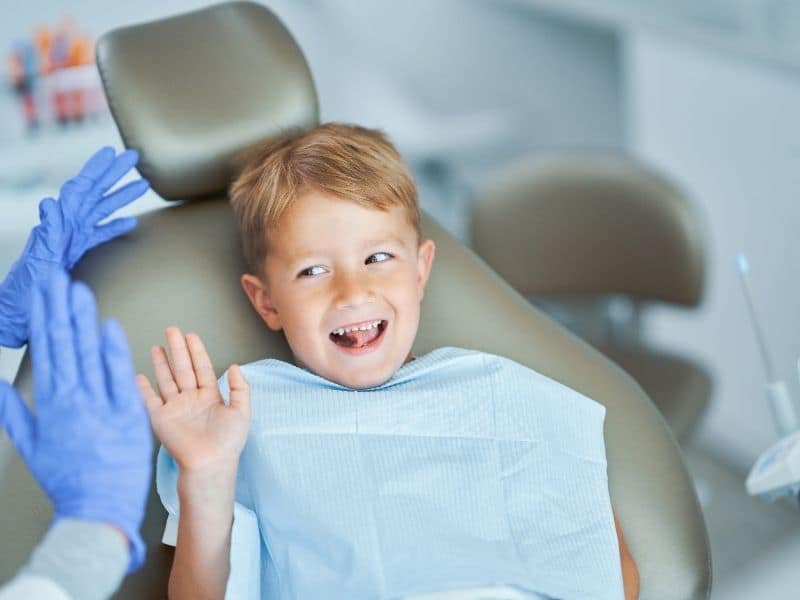 Smiling young child sitting in a dental chair, waving happily while wearing a dental bib during a dental visit.