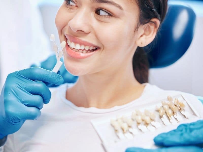 Smiling female patient compares tooth shades with a dental professional using a shade guide during a cosmetic dentistry consultation.
