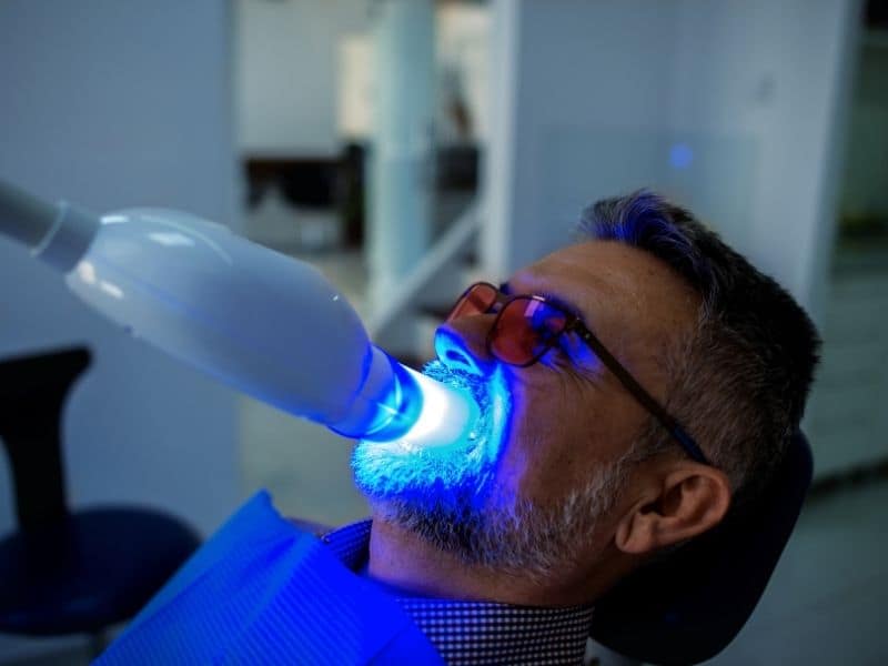 Male patient wearing protective glasses reclines in a dental chair while a blue LED light is used for an in-office teeth whitening treatment.