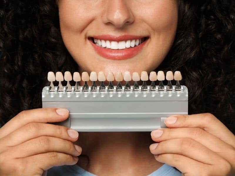 Smiling woman holding a dental shade guide in front of her mouth, displaying a range of tooth color samples used to match and compare natural tooth shades for cosmetic dentistry.